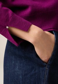Maglione fucsia a maglia con trama a coste, con polsino inclinato e un piccolo strass decorativo. Abbinato a denim blu scuro.