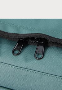 Sac en tissu teal avec une fermeture éclair noire dotée de deux tirettes, présentant une surface texturée et des détails de couture près de l'ouverture.