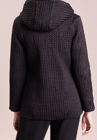 Texturovaná černá bunda s přiléhavým siluetou, vybavená kapucí a výrazným zigzagovým vzorem. Rukávy jsou dlouhé a zúžené.