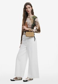 Modella che indossa una camicia a maniche lunghe con un design astratto multicolore, pantaloni bianchi a gamba larga e scarpe beige con dettagli neri, tenendo una borsa intrecciata.