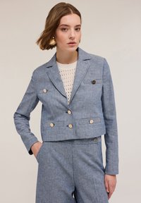Blazer cropped en lin bleu avec un design à double boutonnage, boutons clairs et texture subtile. Associé à un haut en tricot, comportant deux poches latérales.