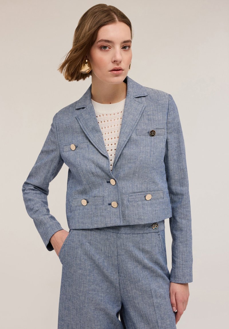 Blazer cropped en lin bleu avec un design à double boutonnage, boutons clairs et texture subtile. Associé à un haut en tricot, comportant deux poches latérales.