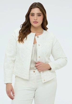 Kobieta o długich falowanych brązowych włosach, ubrana w białą, kwiatową, fakturowaną kurtkę na lekkiej bluzce i białe spodnie, pozująca na jednolitym tle.