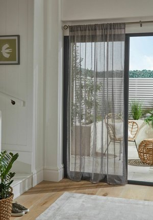 Tenda grigia trasparente appesa sopra una porta scorrevole in vetro che conduce a un patio con piante e mobili in vimini.