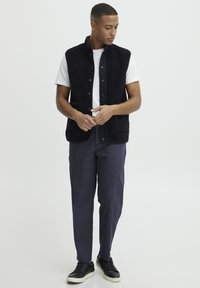 Marine fleece vest med trykknapper, frontlommer, kombinert med en hvit t-skjorte og stripete bukser. Svarte joggesko fullfører antrekket.