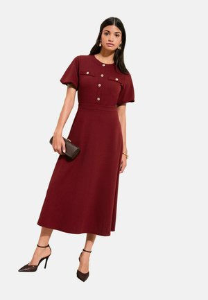 Midi-Kleid in Burgunderrot mit kurzen Puffärmeln, Knopfleiste vorne und zwei Brusttaschen. Gekleidet mit schwarzen Riemchenabsätzen und einer strukturierten Clutch.