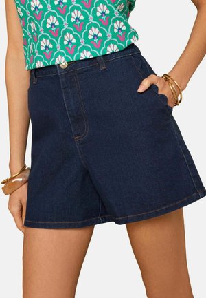 Nő sötétkék farmer rövidnadrágban és zöld mintás felsőben, mindkét csuklóján arany karperecekkel.