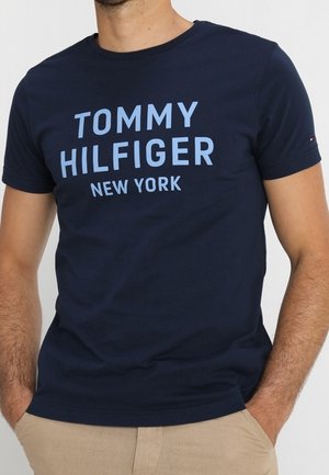 Mann trägt ein dunkelblaues T-Shirt mit kurzen Ärmeln, auf dem in Hellblau „TOMMY HILFIGER NEW YORK“ über der Brust gedruckt ist.