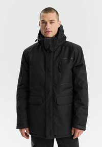 Czarna kurtka na zewnątrz z wysokim kołnierzem, zapinana na zamek, z wieloma kieszeniami. Materiał wydaje się trwały i wodoodporny. Posiada minimalistyczny design.
