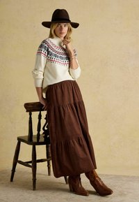 Krémový sveter s červeným a modrým vzorom, hnedá maxi sukňa s volánmi a hnedé kovbojské čižmy. Tmavý plstený klobúk dokončuje celý vzhľad.