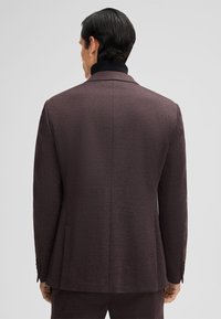Blazer marrone con texture e vestibilità slim, caratterizzato da un colletto nero e cuciture sottili. Include due tasche e quattro bottoni come accenti sulle maniche.