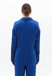 Camicia blu in raso a maniche lunghe con taglio dritto, colletto a notched e vestibilità rilassata. Tessuto liscio con orlo leggermente curvo e polsini arrotolati.