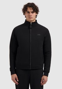 Zwarte ritssluiting sweatshirt gemaakt van zachte stof. Heeft een hoge kraag, zijzakken en een klein logo op de borst. Eenvoudig ontwerp, geen patronen.