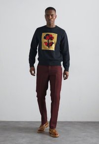 Suéter navy con diseño floral en rojo y amarillo en un parche cuadrado, combinado con pantalones burdeos y zapatos de cuero marrón.
