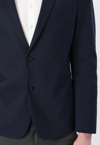 Navy blue blazer made of smooth fabric. Features two button closures, notched lapels, and a chest pocket with a tailored fit.