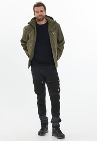 Veste imperméable verte olive pour homme avec capuche, associée à un pantalon cargo noir. Les pantalons sont dotés de poches et de panneaux texturés variés. Bottes de randonnée noires.