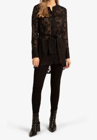 Donna che indossa una blusa nera semi-trasparente a fiori con cintura in vita, leggings neri e stivaletti neri con tacco alto su sfondo bianco.