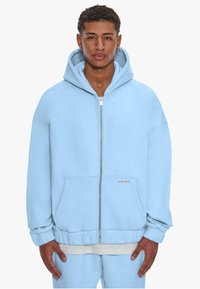 Light blue zip-up hoodie made of soft material. Features a kangaroo pocket, ribbed cuffs, and a spacious hood. Casual oversized fit.