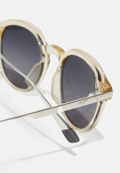 Round sunglasses with a transparent beige frame, dark lenses, and silver metal arms. Notable logo detail on the arm and textured black tips.
