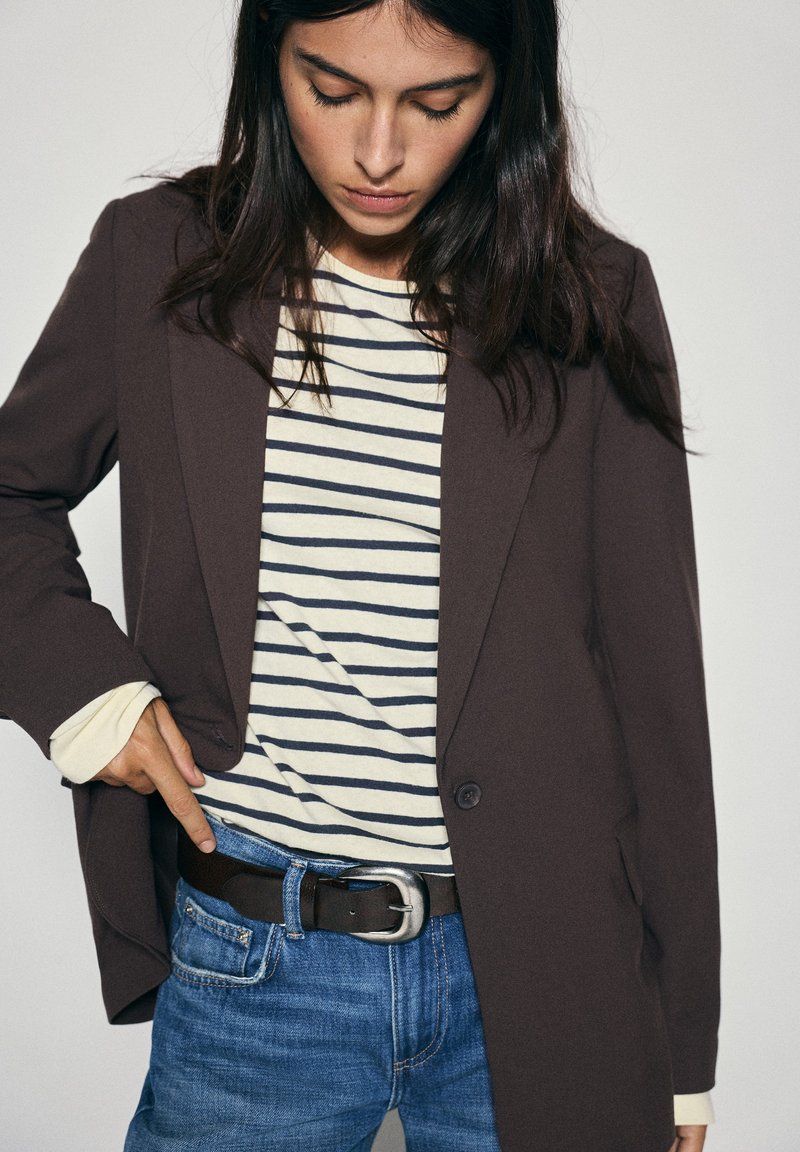 Femme aux cheveux foncés portant un blazer marron sur une chemise rayée, un jean bleu et une ceinture noire avec une boucle argentée, regardant vers le bas sa taille.