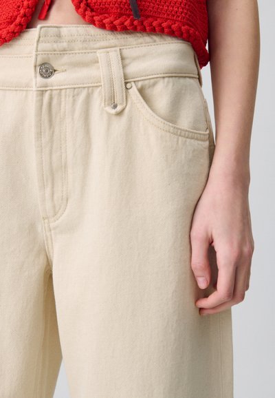 Pantalones de corte amplio en color beige, hechos de algodón, con cintura alta, cierre de botón metálico y bolsillos laterales con una sutil textura acanalada.