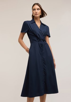 Frau trägt ein marineblaues Midikleid mit Knöpfen, kurzen Ärmeln, Kragen und tailliertem Detail, steht mit einer Hand in der Tasche.