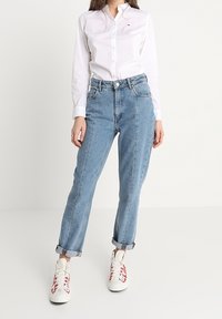Camisa blanca ligera de botones, jeans de mezclilla azul claro de cintura alta arremangados en los puños, y zapatillas blancas con detalles rojos.