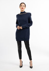 Pulover oversized de culoare bleumarin cu guler înalt, detalii cu lanțuri aurii și mâneci lungi, asortat cu pantaloni de piele negri, ajustați și pantofi cu toc negri.