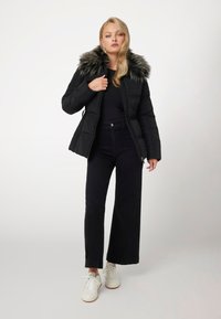 Juodas pūkuotas švarkas su dirbtiniu kailiniu apykakle, užtrauktuku ir šoninėmis kišenėmis, dėvimas ant juodos palaidinės, derinamas su plačiomis tamsiomis džinsomis ir baltomis sportinėmis bateliais.