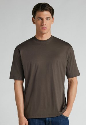 Junger Mann mit kurzen dunklen Haaren, trägt ein schlichtes dunkelbraunes kurzärmeliges T-Shirt und blaue Jeans, steht vor einem grauen Hintergrund.