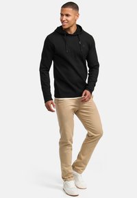 Sudadera negra acanalada con un bolsillo tipo canguro, combinada con chinos beige claros y zapatillas blancas. La capucha cuenta con cordones y un sutil detalle del logo.