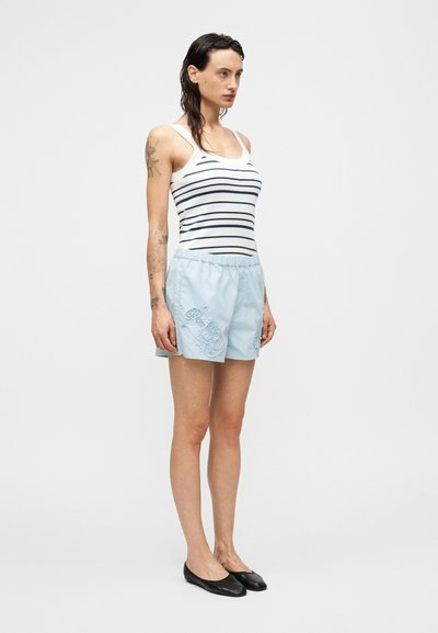 Ανοιχτό μπλε σορτς με ελαστική μέση, διακοσμημένο με κεντητά σχέδια στο μπροστινό μέρος. Συνδυάζεται με ένα μπλε ναυτικό και λευκό ριγέ αμάνικο τοπ.