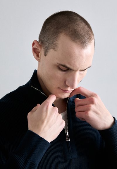 Marineblaues gestreiftes Pullover mit einem Reißverschlusskragen. Das Modell zieht den Reißverschluss mit beiden Händen nach oben und zeigt eine leichte Innenschicht darunter.