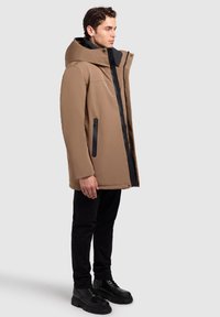 Brun, huvad, vattentät jacka med svart dragkedja och detaljer. Har två sidofickor med dragkedja och en avslappnad passform. Bärs över svarta byxor och stövlar.