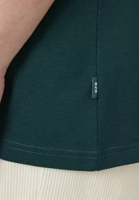 T-shirt vert foncé avec une texture lisse, présentant une petite étiquette latérale portant la mention "BZB" et un ourlet droit. Un pantalon de couleur claire est visible en dessous.