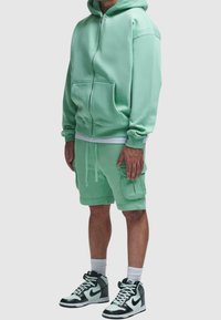Homme portant un sweat à capuche zippé vert menthe et un short cargo assorti, avec des baskets montantes Nike noires et blanches et des chaussettes blanches.