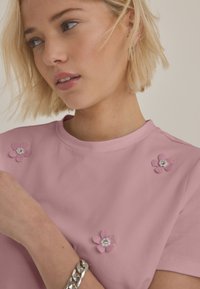 Roza majica s kratkimi rokavi in tremi cvetličnimi aplikacijami, ki imajo teksturirane cvetne liste in okraske v sredini. Gladek material in zaobljen izrez na vratu.
