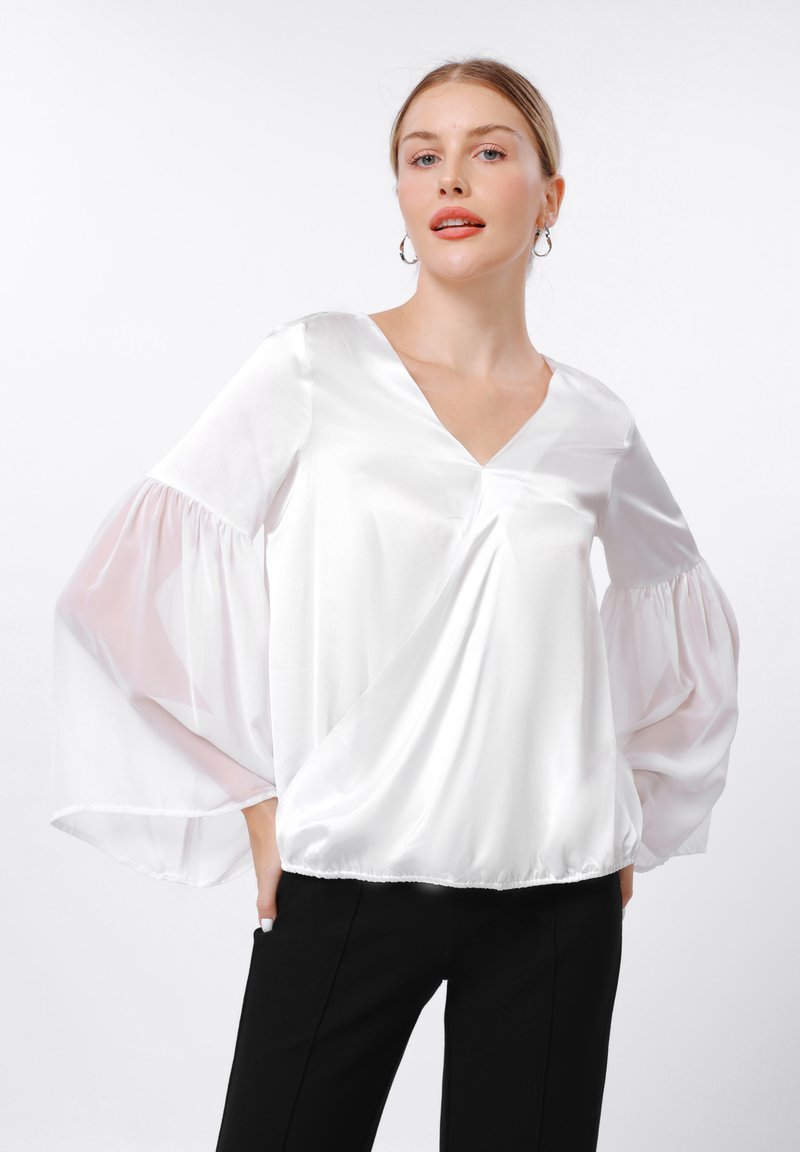 Blouse en satin blanc avec un col en V, des manches légères et bouffantes, et un ourlet froncé, associée à un pantalon noir pour le contraste.