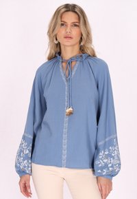 Blusa azzurra chiara con maniche lunghe a palloncino, decorata con motivi floreali ricamati in bianco e una scollatura a laccio con un tassello decorativo.