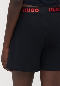 Svarta bomullsshorts har ett rött elastiskt midjeband med "HUGO"-logga. Designen är enkel med en slät textur och rena linjer.