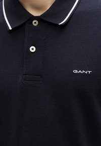 Polo bleu marine en tissu texturé, col rayé blanc, patte de boutonnage à deux boutons et logo "GANT" brodé sur la poitrine.