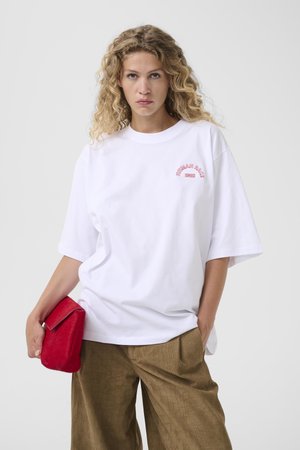 Blonde vrouw met krullend haar draagt een wit oversized T-shirt en bruine textuurbroek, houdt een rode clutch vast tegen een effen achtergrond.