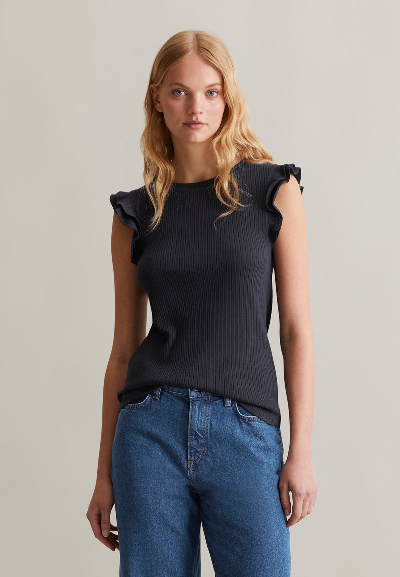 Zwarte geribbelde gebreide top met ruches op de schouders, gecombineerd met blauwe rechte denim jeans. Eenvoudig, textuurvol ontwerp dat de silhouet benadrukt.
