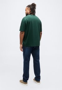 Άνδρας με ντραντλοκς, ντυμένος με σκούρο πράσινο μπλουζάκι, μπλε τζιν και μπεζ αθλητικά παπούτσια, στέκεται γυρισμένος με την πλάτη σε απλό λευκό φόντο.