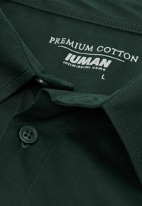 Camisa de botón de color verde oscuro con cuello, con un botón negro visible y una etiqueta blanca impresa que dice "Algodón Premium" y talla "L".