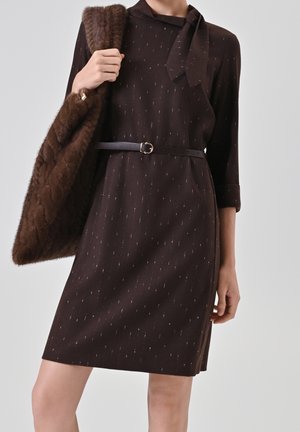 Woman wearing a dark brown patterned dress with a thin belt, holding a large brown fur tote bag over her shoulder.