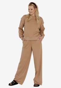 Beżowy zestaw bluzki i spodni z szerokimi nogawkami, wykonany z miękkiego materiału, o luźnym kroju i prążkowanych mankietach. Czarne sandały na paski.