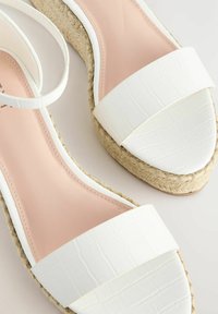 Valged espadrillid sandaalid on varustatud tekstureeritud krokodillimustriga varvaste rihmaga, pehme roosa sisetallaga ja kootud jute kesktaldaga.