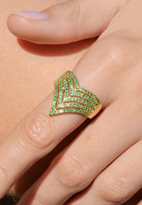 Bague en ton or présentant un design en chevron avec plusieurs rangées de pierres précieuses vertes pour la texture et l'éclat, posée sur une main nue.