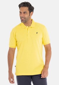 Polo shirt jaune en coton, avec un col et des manches courtes. Emblème noir sur la poitrine. Design simple avec une texture lisse.
